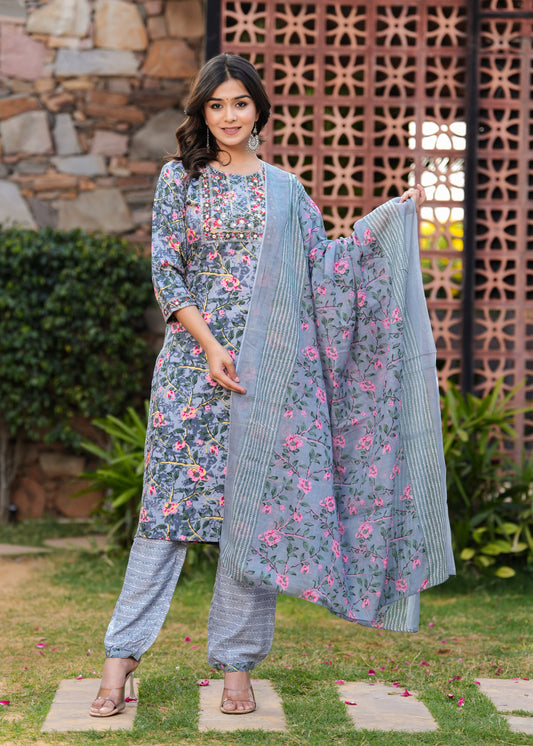 Woman in floral dress with matching dupatta standing outdoors against a stone wall.