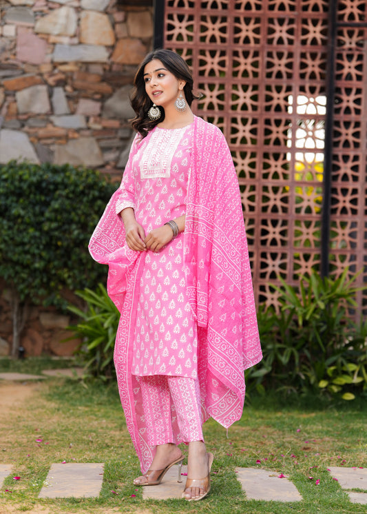 Woman in a pink traditional outfit standing outdoors with a stone wall and plants in the background