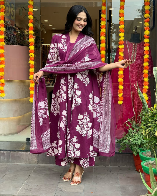 Woman in a purple traditional outfit with white patterns standing in front of a decorated entrance.