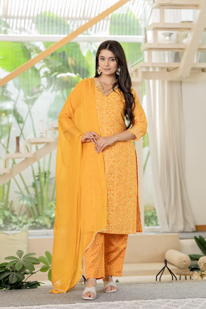 Woman in a yellow traditional outfit standing in a modern indoor setting with plants and a staircase.