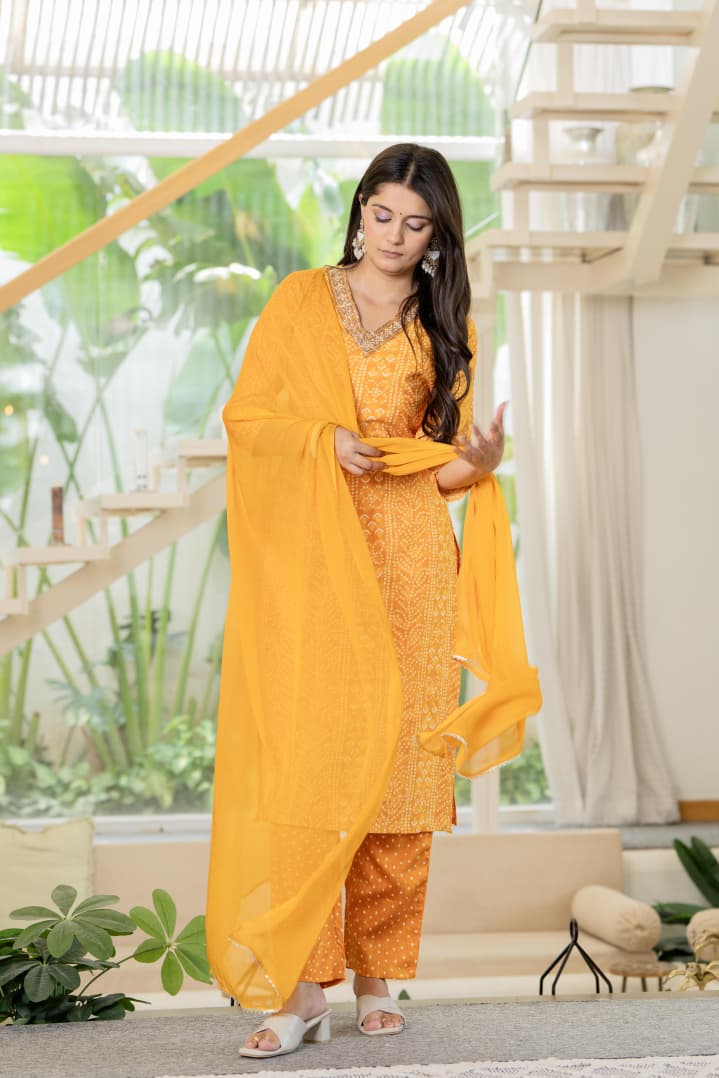 Woman in a yellow traditional outfit standing in a modern indoor setting with plants and a staircase.