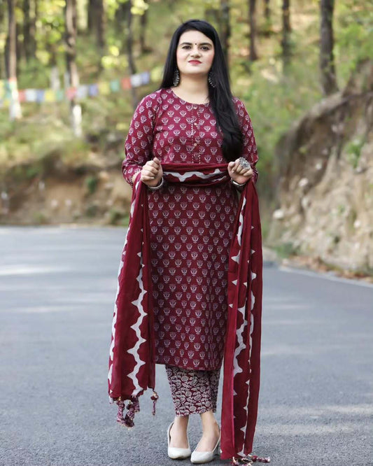 Woman in a maroon traditional outfit with a patterned scarf standing on a road with trees in the background.
