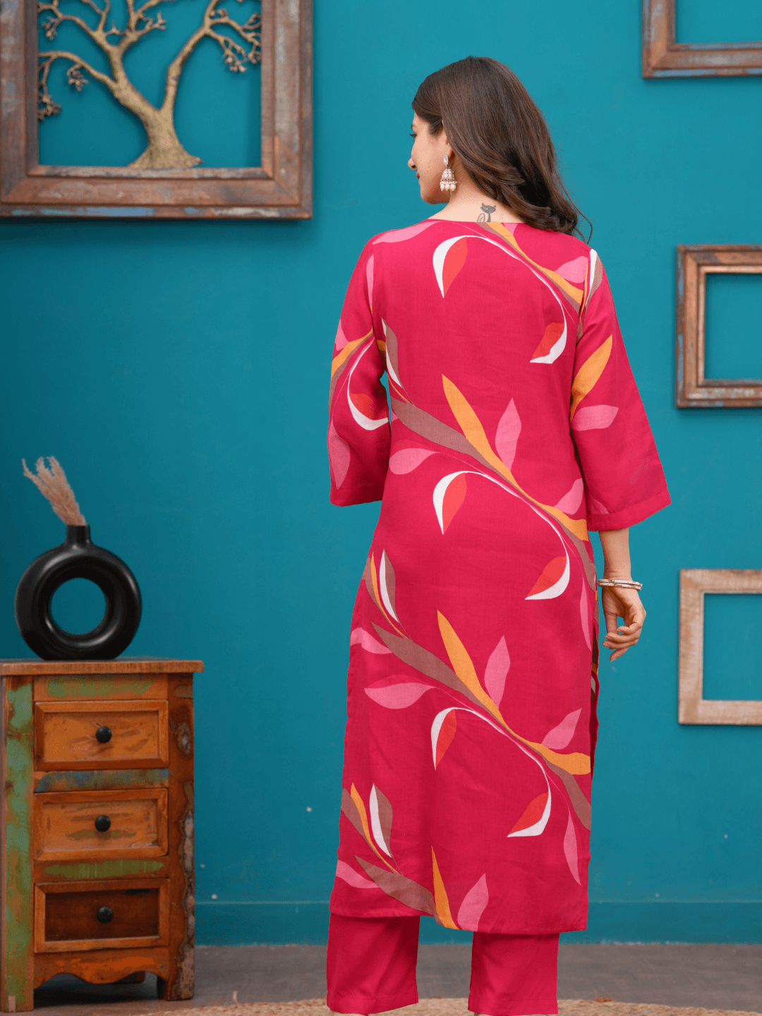 Woman wearing a pink patterned outfit against a teal wall with wooden furniture.