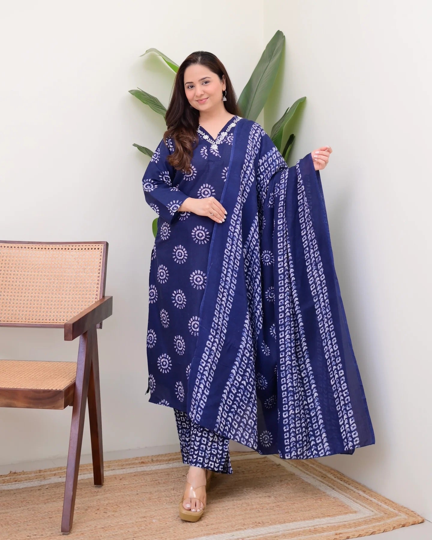 Woman wearing a blue and white patterned outfit standing next to a wooden chair with a plant in the background.