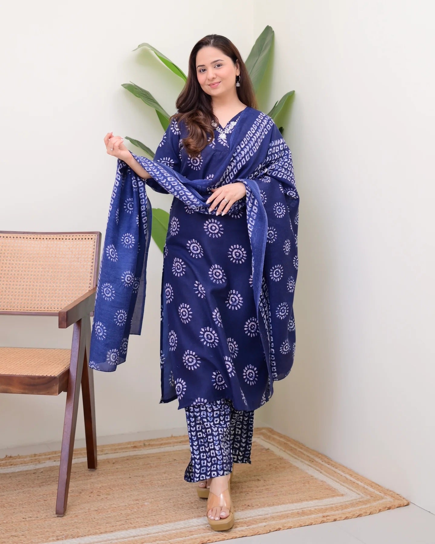 Woman holding a blue patterned scarf in a room with a chair and plants.