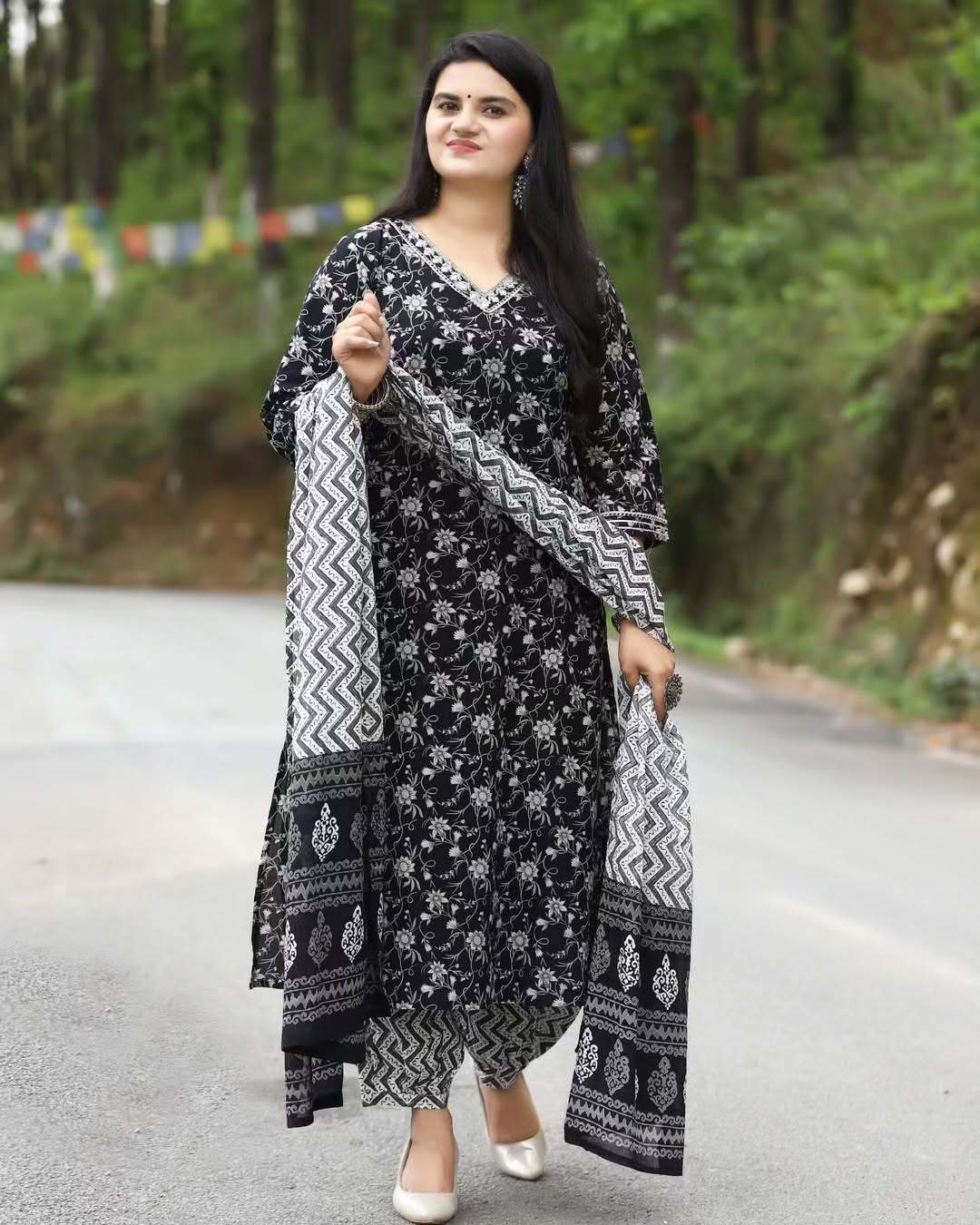 Woman in a black floral dress with a patterned dupatta standing on a road with greenery in the background
