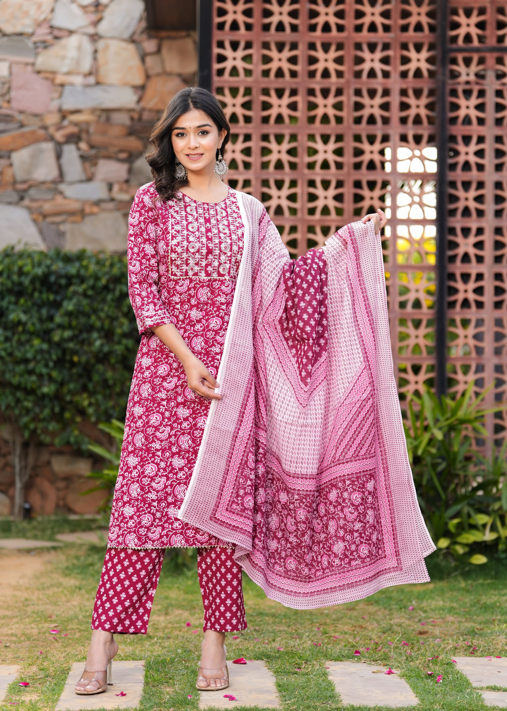 Woman in a pink and white traditional outfit standing outdoors with a decorative wall and plants in the background.