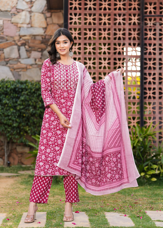 Woman in a pink and white traditional outfit standing outdoors with a decorative wall and plants in the background.