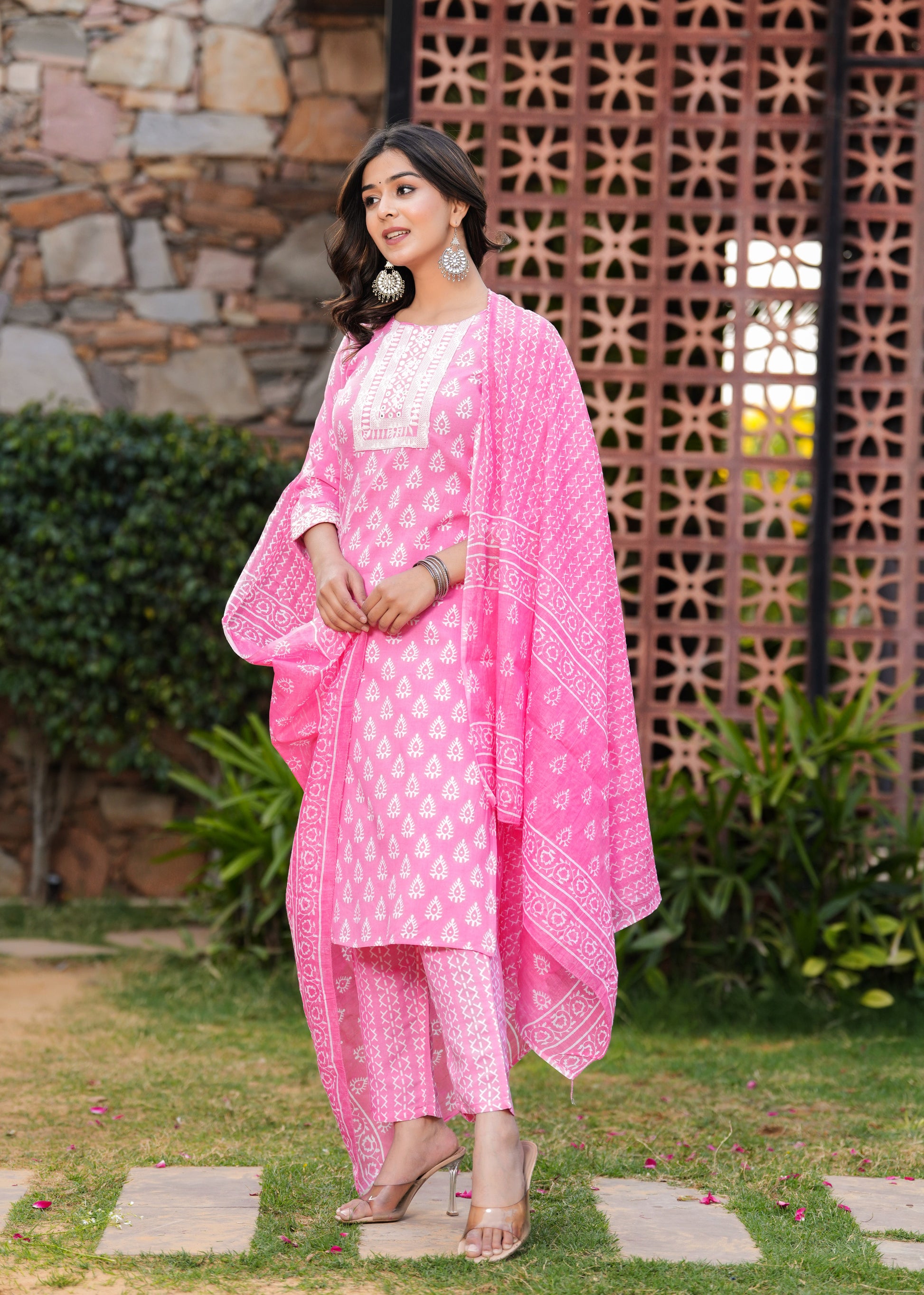Woman in a pink traditional outfit standing outdoors with a stone wall and plants in the background