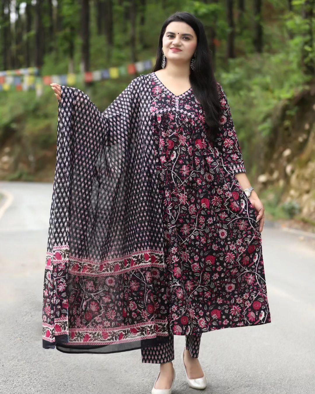 Woman holding a black and white patterned dupatta with a floral dress on a road with greenery in the background